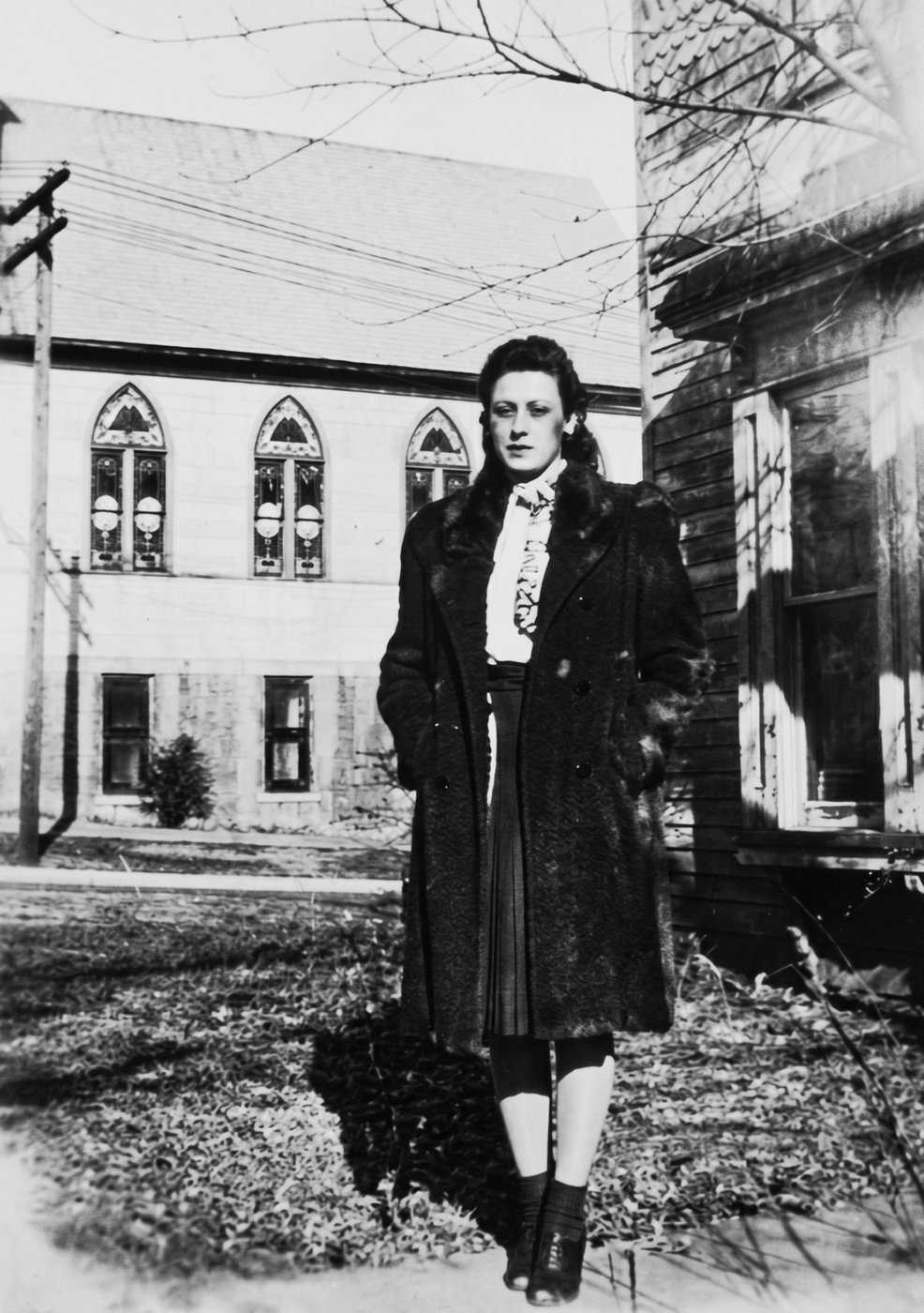 Norma in a long fur coat standing on a sidewalk in front of a church